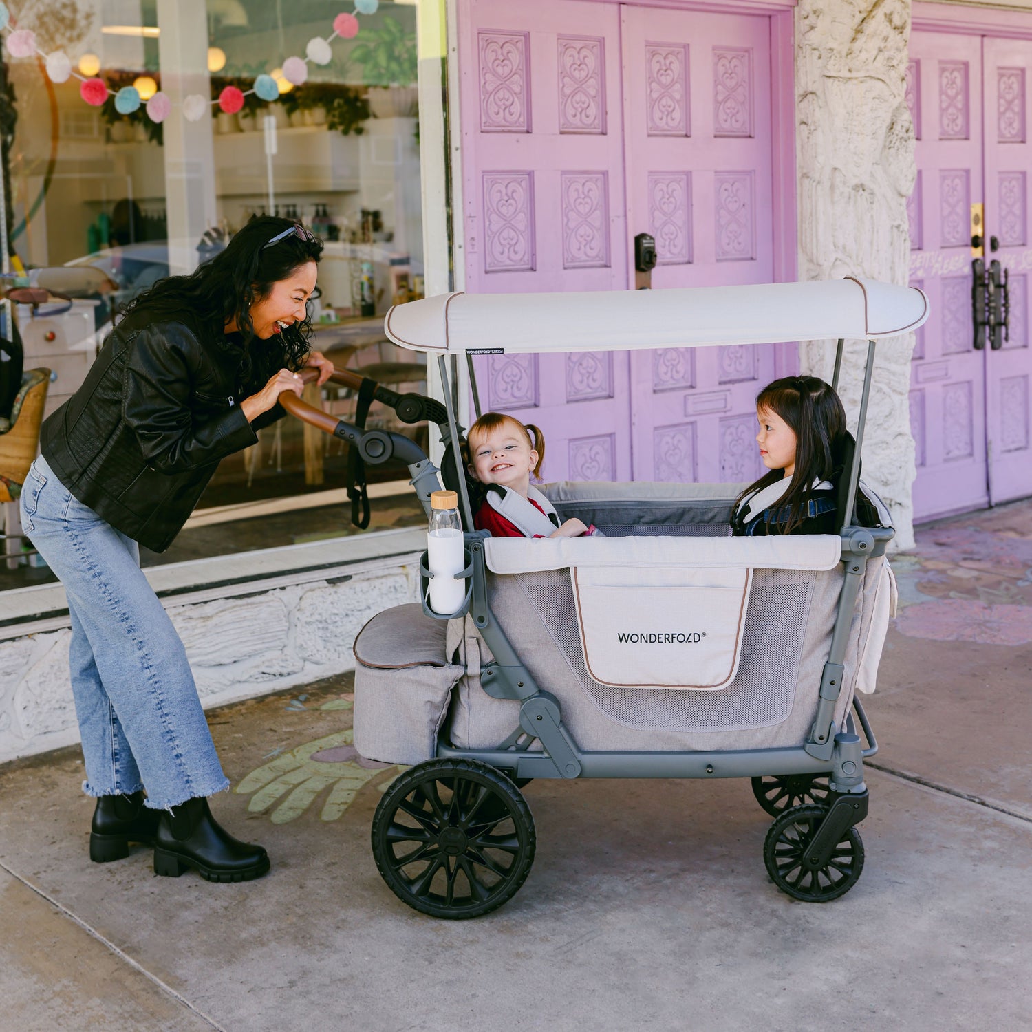 A woman joyfully pushing a WonderFold L2 stroller wagon with two children inside, featuring a canopy and stylish design in front of a vibrant storefront.