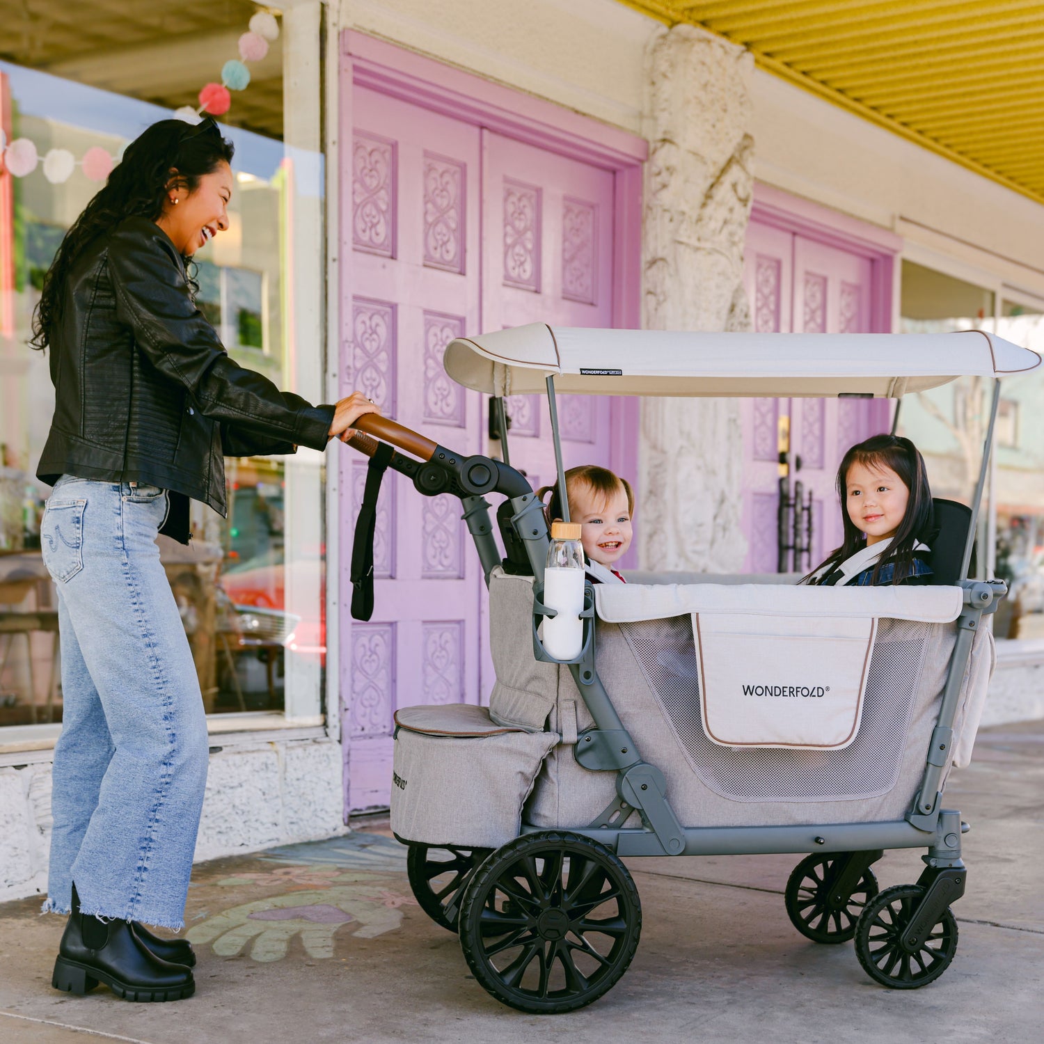 A woman pushing the WonderFold L2 stroller wagon with two happy children inside, strolling past a colorful storefront.