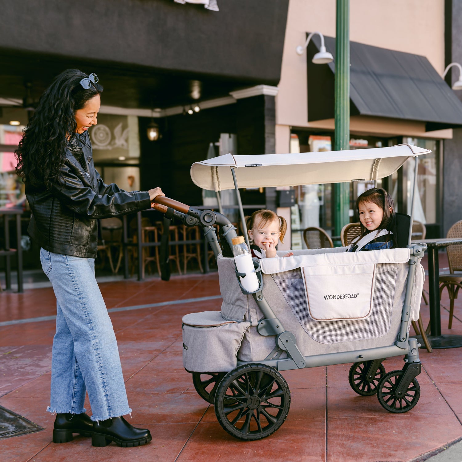 A woman pushing the WonderFold L2 stroller wagon with two children inside, enjoying a walk through a city area.