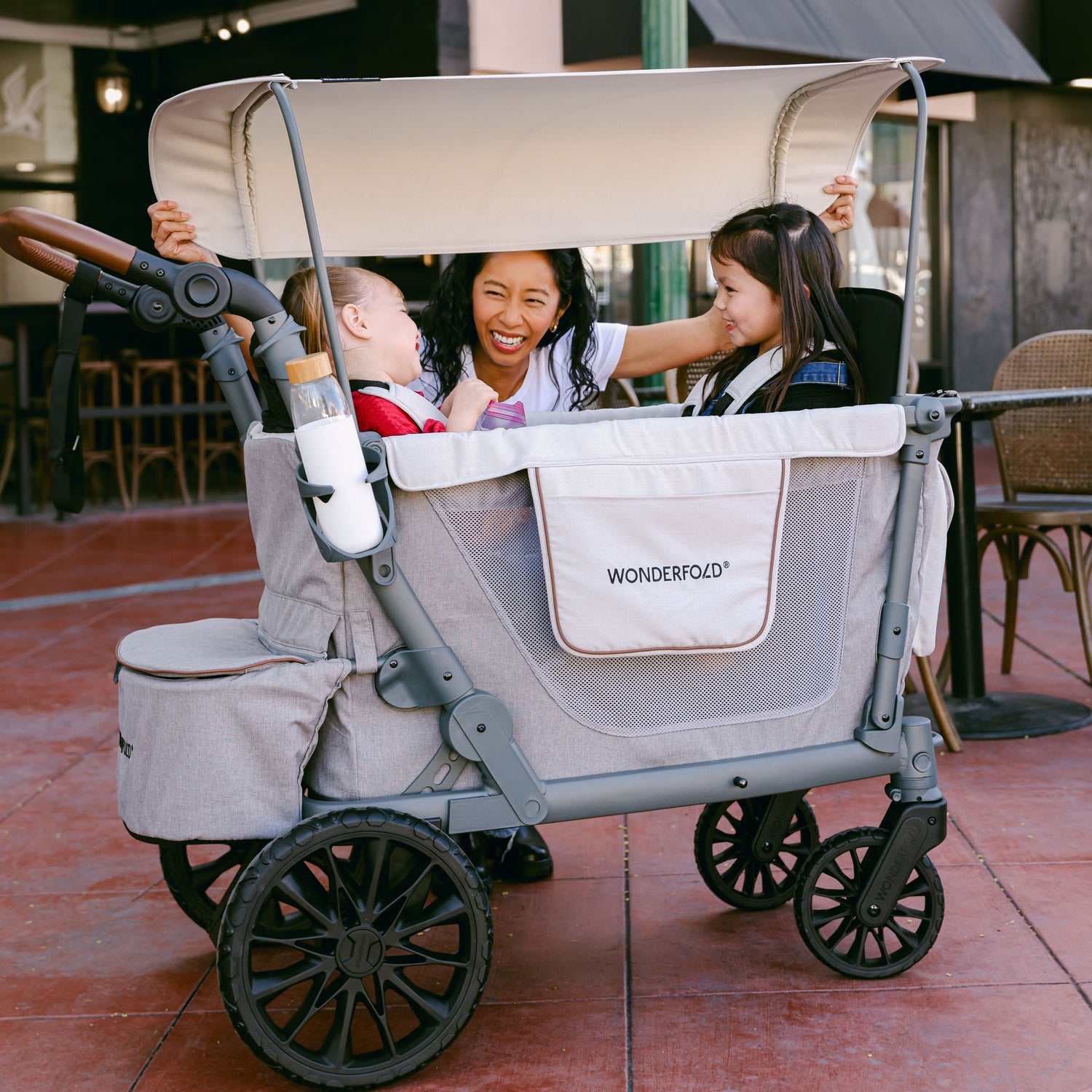 A woman adjusting the canopy shade of the WonderFold L2 stroller wagon while interacting with two smiling children inside.