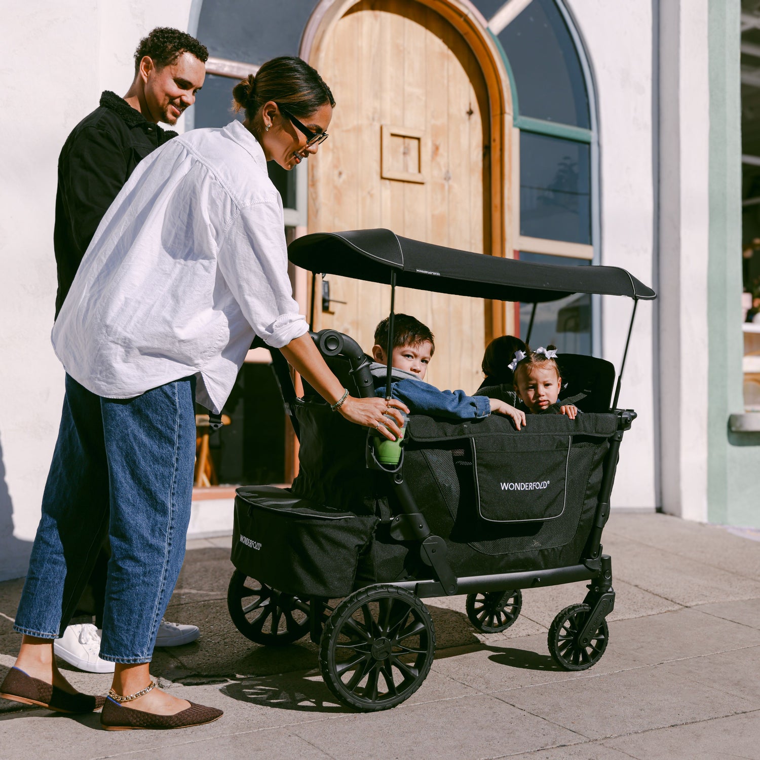 A woman pushing the WonderFold L4 stroller wagon with four children inside, strolling near a café.