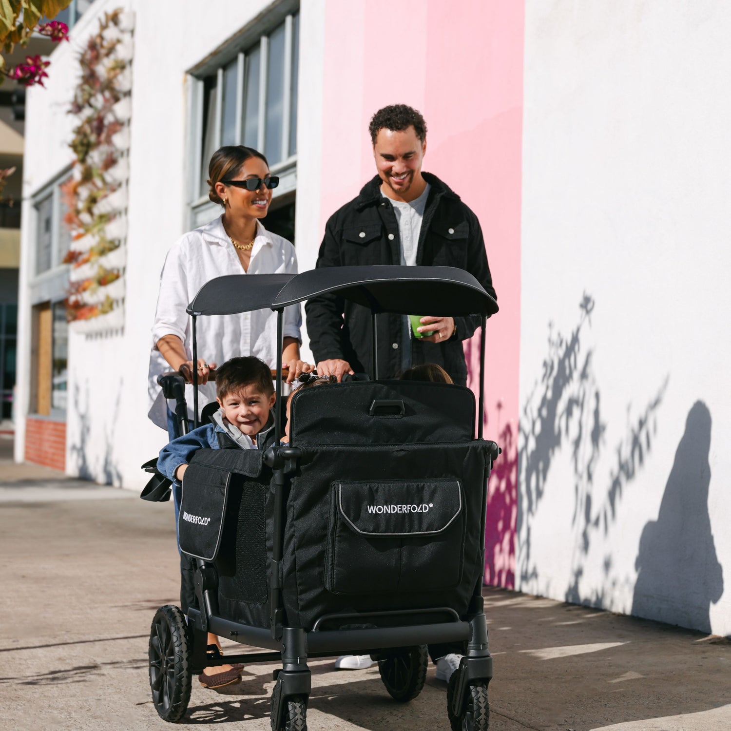 A family of three strolling with the WonderFold L4 stroller wagon, featuring a black canopy and spacious seating.