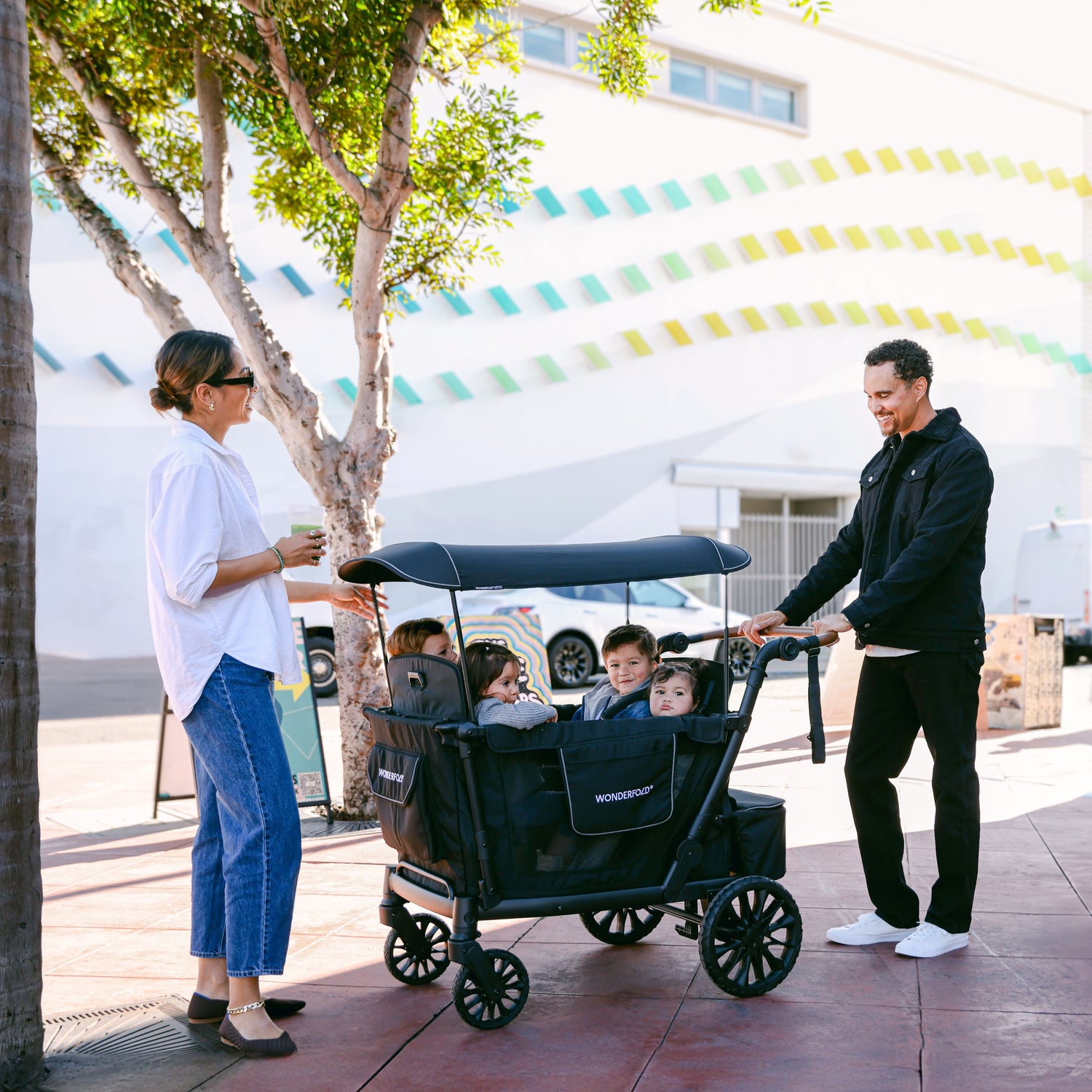 A father pushing the WonderFold L4 stroller wagon with four children inside, while the mother enjoys a conversation nearby.