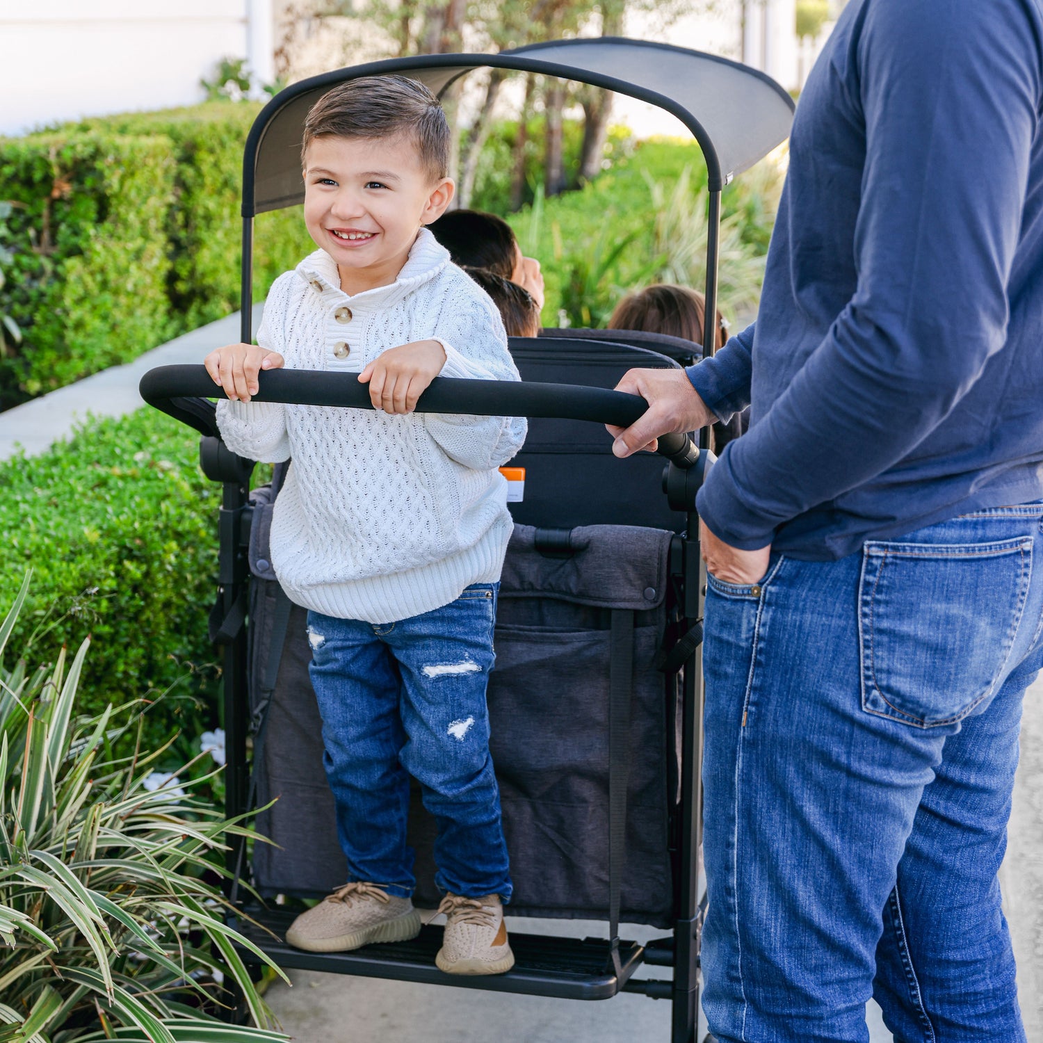 Child standing on the Buddy Board accessory, securely attached to a WonderFold W4 stroller wagon